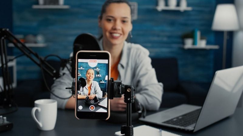 Mulher sorridente em home office gravando conteúdo para redes sociais, com o celular exibindo uma pessoa ao vivo e iluminação de estúdio ao lado do computador, tema sobre exposição do trabalhador nas redes sociais