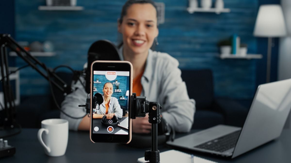 Mulher sorridente em home office gravando conteúdo para redes sociais, com o celular exibindo uma pessoa ao vivo e iluminação de estúdio ao lado do computador, tema sobre exposição do trabalhador nas redes sociais
