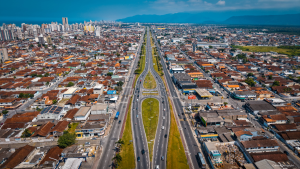 Vista aérea de uma avenida principal entre bairros residenciais, com ruas movimentadas e telhados de casas, ao fundo serras e céu azul.