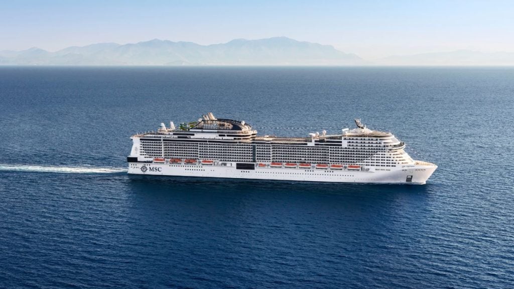 MSC Cruzeiros em Santos: MSC Virtuosa, navio de cruzeiro moderno da MSC Cruzeiros, navegando no mar em uma imagem aérea com fundo de montanhas e céu claro.