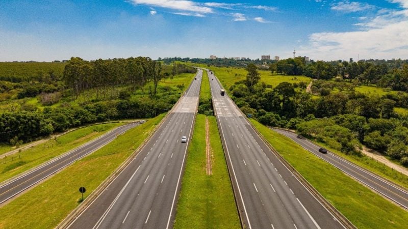 Movimento nas rodovias durante feriado de 7 de setembro: Vista aérea de uma rodovia com faixas livres e veículos em movimento, cercada por áreas verdes e árvores.