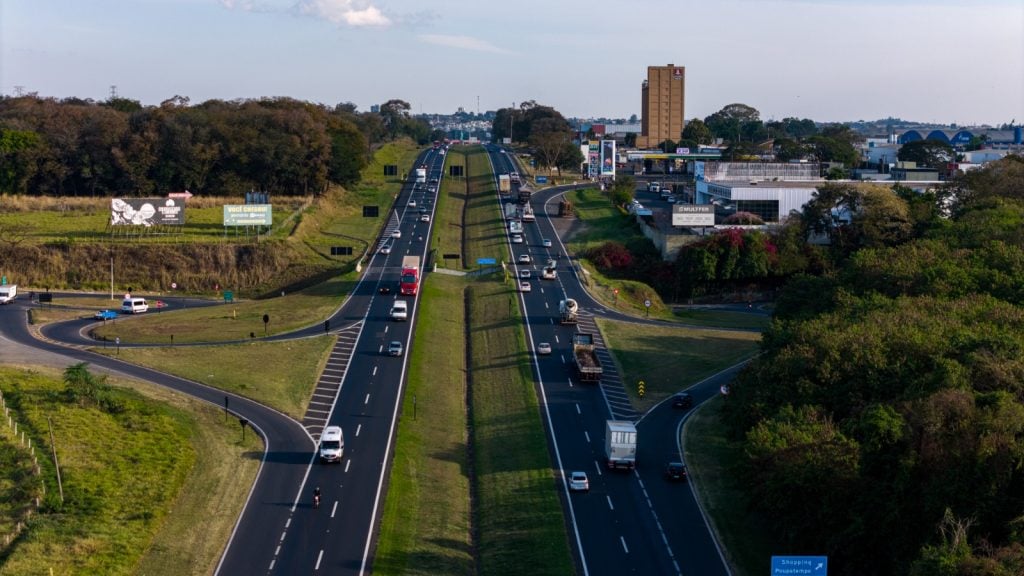 Movimento nas rodovias durante feriado de 7 de setembro: vista aérea de uma rodovia com veículos em fluxo, faixas bem sinalizadas e retorno à frente em meio a área verde.