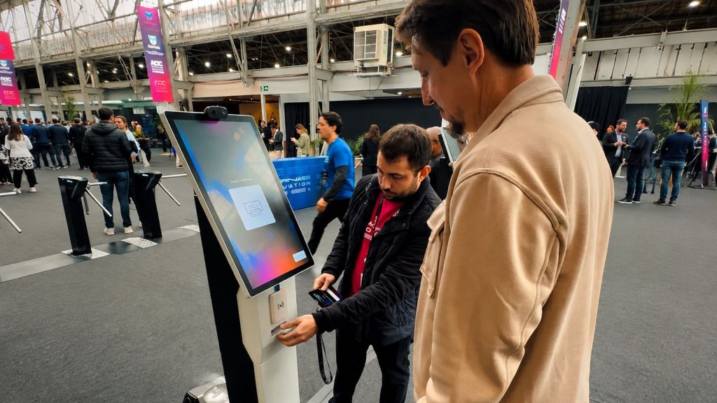 Participante usando um totem de autoatendimento com tela e leitor no evento Campinas Innovation Week, com foco em reconhecimento facial, RFID e Inteligência Artificial para gerar dados estratégicos.