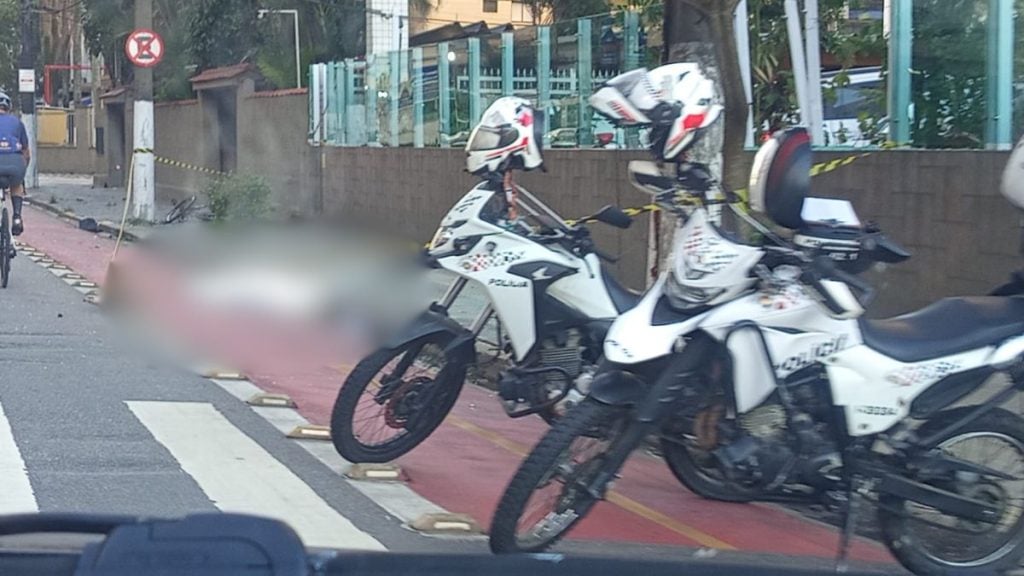 Ciclista atropelado em via urbana, com motocicleta e faixas de ciclovia ao fundo; veiculo parece ter invadido a área enquanto o trânsito é observado.