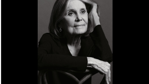 Retrato em preto e branco de Gloria Steinem em estúdio, com cabelo longo e expressão serena, apoiando a cabeça na mão e os braços cruzados em pose clássica.