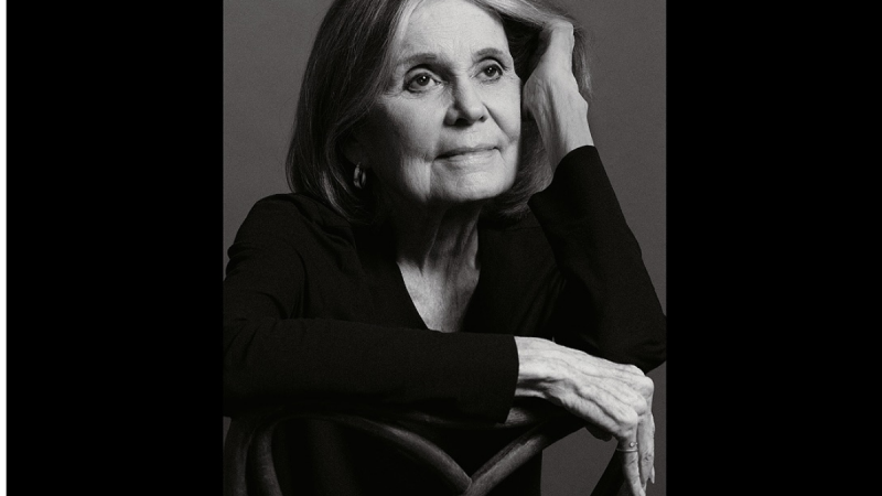 Retrato em preto e branco de Gloria Steinem em estúdio, com cabelo longo e expressão serena, apoiando a cabeça na mão e os braços cruzados em pose clássica.