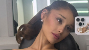 Ariana Grande sorrindo em selfie no escritório, com cabelo preso e maquiagem leve, usando celular com capa decorada, em ambiente de trabalho com janelas ao fundo.