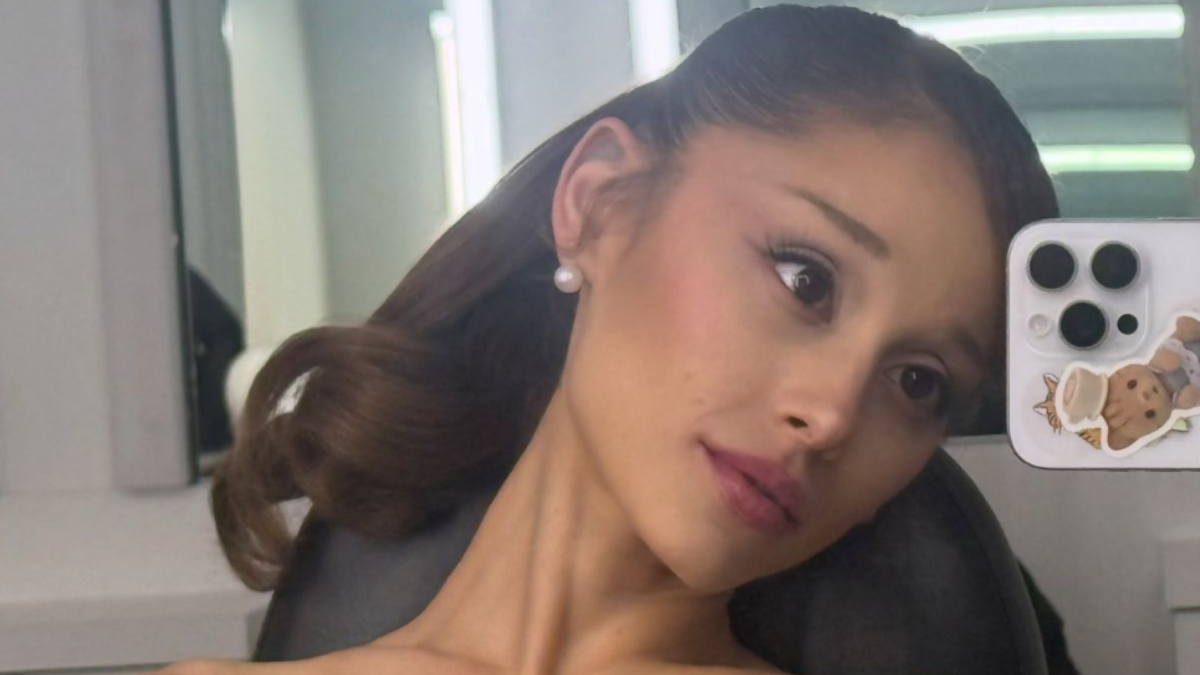 Ariana Grande sorrindo em selfie no escritório, com cabelo preso e maquiagem leve, usando celular com capa decorada, em ambiente de trabalho com janelas ao fundo.