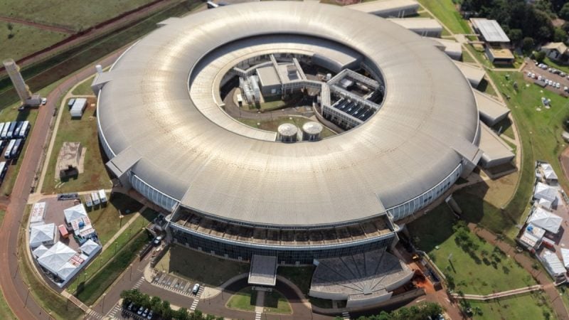 Vista aérea do Sirius em Campinas, com o grande edifício circular e estruturas de pesquisa e tecnologia ao redor, em cenário urbano e verde.