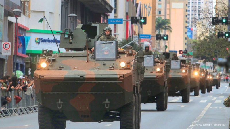 Militares em veículos blindados desfilam em uma avenida durante o 7 de setembro em Campinas, com público nas laterais e sinalização de trânsito ao fundo.