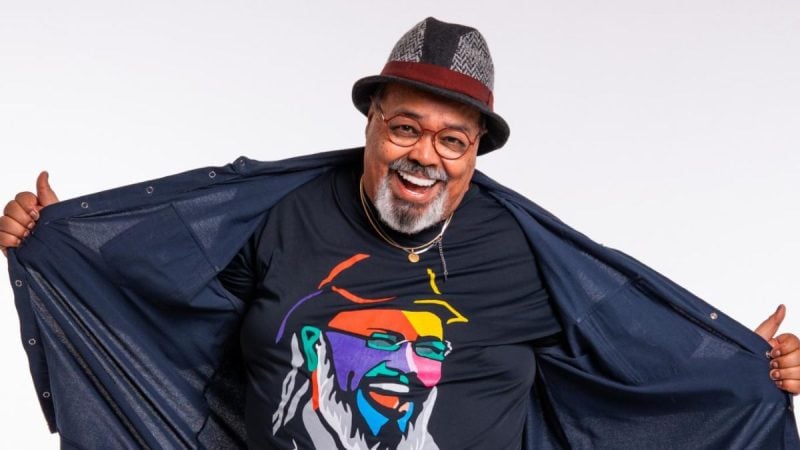 Jorge Aragão em foto sorrindo, usando chapéu e óculos, com a mão aberta em clima descontraído, durante divulgação de sua apresentação na festa de lançamento do Carnaval de Santos 2027.