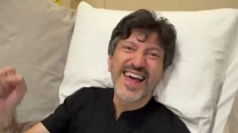Lito Souza sorrindo e rindo sentado em uma cama, com travesseiros claros ao fundo, em ambiente doméstico, olhando para a câmera.