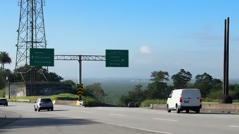 Rodovia em estrada com placas verdes de orientação, veículos trafegando e vista de região montanhosa ao fundo sob céu azul.