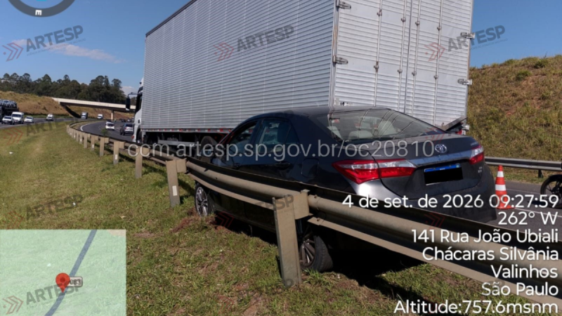 Carro escuro parado em acostamento ao lado de um caminhão em rodovia, com cones de sinalização e placa de localização; imagem com marcações de data e hora na tela.