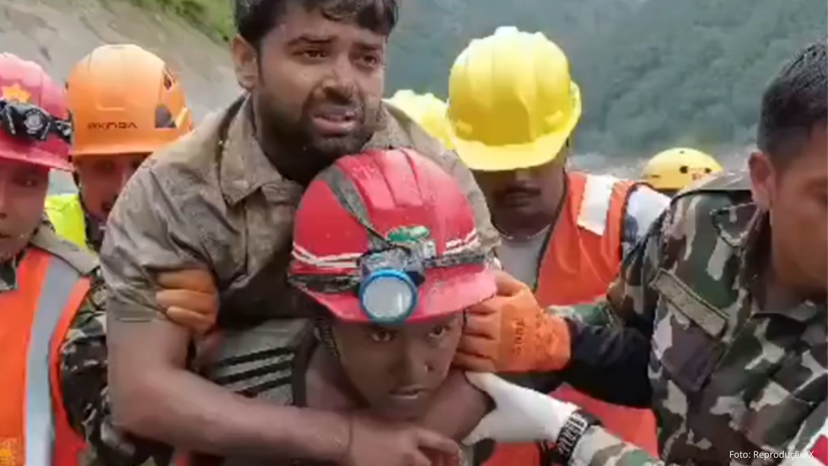 Homem em primeiro plano ajuda trabalhador em meio a equipes de resgate, com coletes laranja e capacetes em um ambiente externo. A cena transmite socorro e cuidado após um acidente.