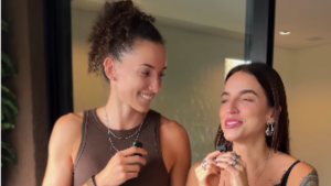 Duas mulheres sorrindo e conversando em um ambiente interno, com uma segurando um colar e a outra usando brincos e maquiagem leve, luz natural suave.