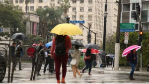 Pessoas caminhando em rua movimentada durante a chuva, usando guarda-chuvas coloridos como amarelo, azul e rosa, com prédios ao fundo e semáforos na cidade.