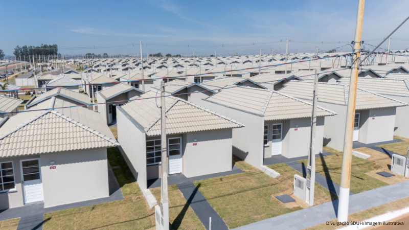 Vista aérea de um conjunto habitacional com casas térreas modernas e telhados brancos em bairro planejado, em dia de céu azul.
