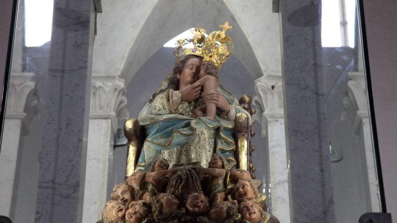 Nossa Senhora do Monte Serrat em imagem devocional, entronizada com o Menino Jesus nos braços, cercada por anjos e detalhes dourados em um altar de igreja.