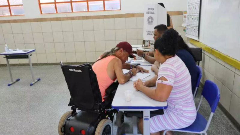 Fotografia em ambiente interno de uma seção eleitoral, mostrando uma pessoa com deficiência física sentada em uma cadeira de rodas motorizada preta (com a marca "Ottobock" visível no encosto) enquanto assina documentos em uma mesa escolar azul e branca.