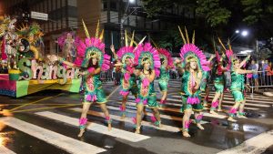 Retorno do desfile das escolas de samba em Campinas em 2027: ala de fantasias coloridas com integrantes dançando na passarela à noite, com carros alegóricos e bandeiras.