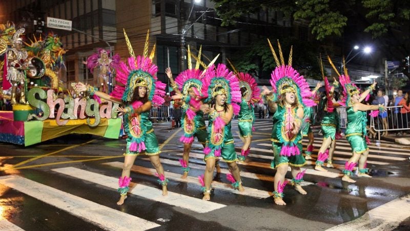 Retorno do desfile das escolas de samba em Campinas em 2027: ala de fantasias coloridas com integrantes dançando na passarela à noite, com carros alegóricos e bandeiras.