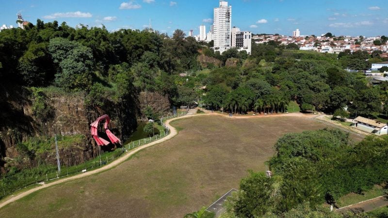 Vista aérea da Pedreira do Chapadão em Campinas, com área gramada e trilha entre vegetação densa, céu com nuvens e cidade ao fundo, cenário do Cine Autorama cancelado pela previsão de chuva.