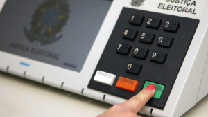 Teclado de uma urna eletrônica com botões de números e opções como BRANCO, CORRIGE e CORRIGE, com dedo apontando para o botão VERDE de confirmação, em cenário de Justiça Eleitoral.