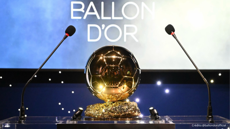 Bola de futebol dourada com brilho sobre um pedestal em evento, com microfones direcionados para o centro e o texto “BALLON D’OR” ao fundo.