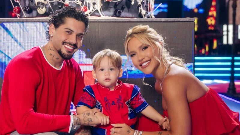 Virginia e Zé Felipe posam sorridentes ao lado do filho durante festa, em ambiente de comemoração com decoração e luzes ao fundo.