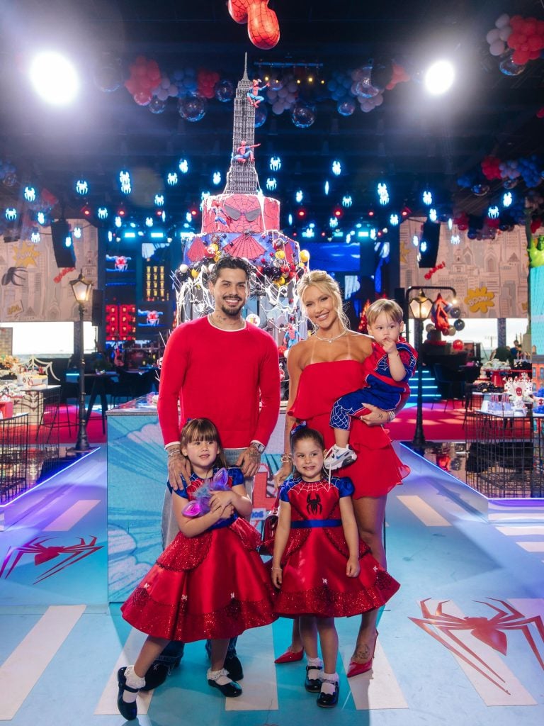 Virginia e Zé Felipe posam sorridentes ao lado do filho durante festa, em ambiente de comemoração com decoração e luzes ao fundo.
