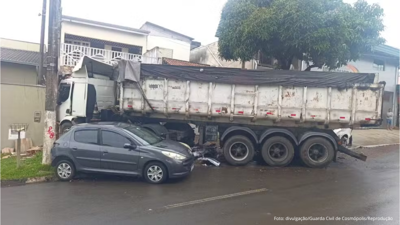 Caminhão de carga com compartimento basculante tombado na via e um carro pequeno estacionado ao lado, em área urbana, com vegetação e prédios ao fundo.
