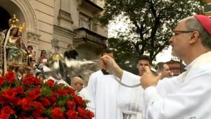 Imagem padre em procissão ao ar livre, segurando o incenso enquanto imagem de Nossa Senhora do Monte Serrat aparece ao fundo, com flores vermelhas em primeiro plano.