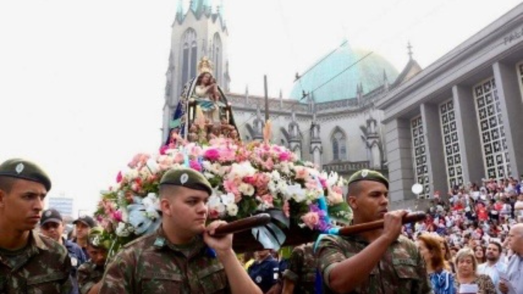 Militares em desfile seguram uma grande coroa de flores durante cerimônia em frente a uma igreja, com multidão acompanhando em um espaço urbano.