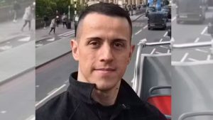 Michel Serra Begali em foto ao ar livre, em uma rua movimentada, ao lado de um veículo, com expressão neutra e fundo urbano desfocado