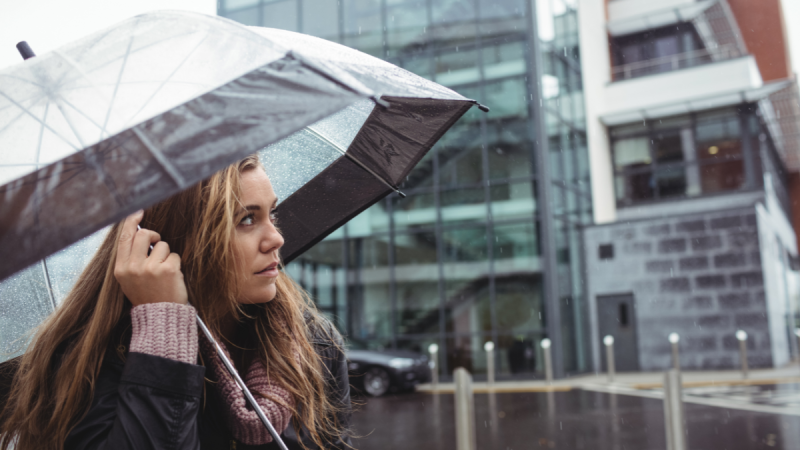 Mulher com guarda-chuva na chuva em frente a um prédio moderno, segurando o cabo com expressão de atenção, em cenário urbano.