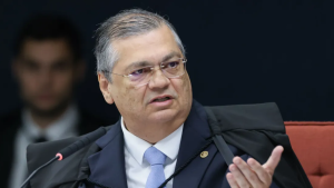 Flávio Dino sentado em uma reunião e falando ao lado de microfones, com expressão séria. Fundo escuro e cadeira vermelha.
