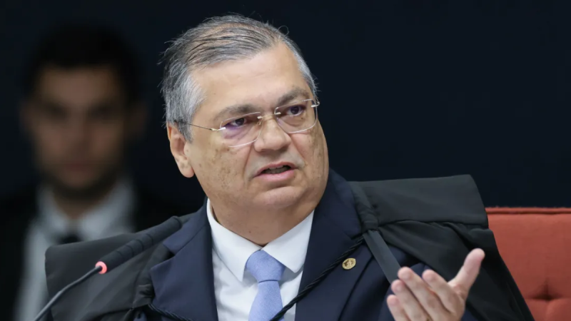Flávio Dino sentado em uma reunião e falando ao lado de microfones, com expressão séria. Fundo escuro e cadeira vermelha.