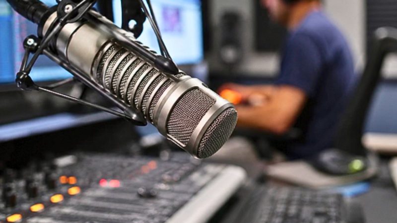 Microfone profissional em estúdio de rádio com locutor ao fundo, reforçando o streaming de rádio durante a propaganda eleitoral e ampliando a audiência.