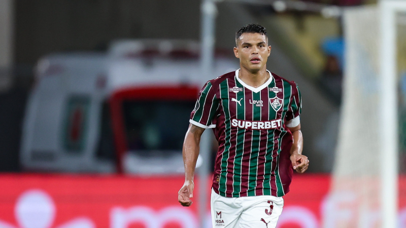 Thiago Silva em campo durante partida pelo Fluminense, com camisa listrada verde e vermelha, em imagem esportiva ao fundo com estádio desfocado; Thiago Silva Ancelotti em destaque na matéria.