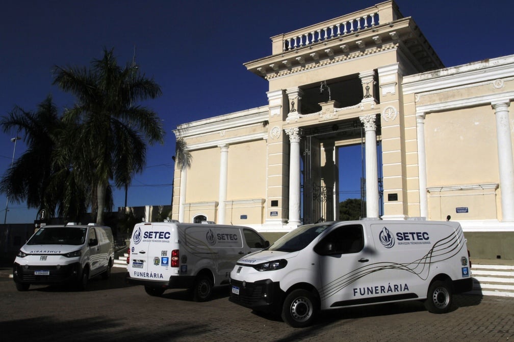 concursos setec: Furgões da SETEC com a palavra FUNERÁRIA estacionados em frente a um prédio histórico sob céu azul, em contexto de divulgação do concurso setec com 40 vagas.
