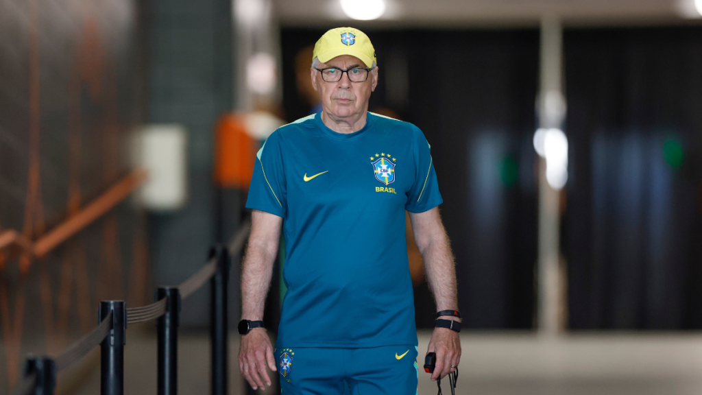 Carlo Ancelotti em foto como técnico da Seleção Brasileira, usando uniforme azul e boné amarelo, em ambiente interno com foco no treinador.