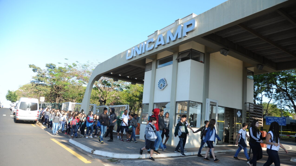 Pessoas em fila caminham em frente a um prédio industrial com a placa “UNICAMP”, em área externa com árvores ao fundo e veículos estacionados na via.