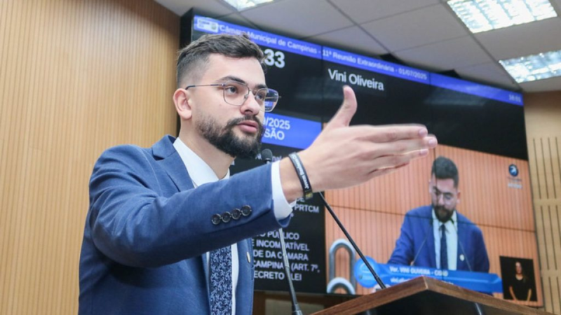 Vini Oliveira é cassado: homem com terno e barba discursa na Câmara de Campinas, apontando para a plateia, enquanto ao fundo aparece outra pessoa na mesa de votação