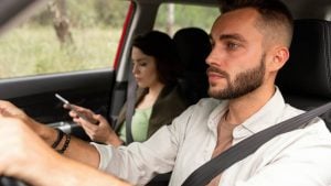 Homem dirigindo um carro, com uma jovem no banco de trás usando o celular durante uma viagem, tema sobre Uber e possibilidade de vídeo ao vivo em deslocamentos