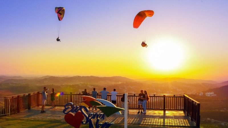 Pessoas voando de parapente na Serra Negra ao pôr do sol, com parapentes coloridos no céu e uma área de decolagem em primeiro plano.