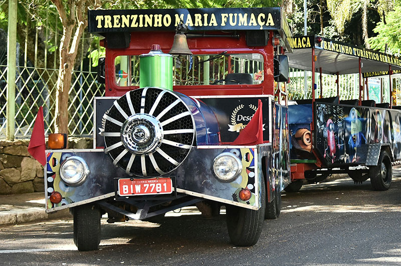 Trenzinho de Serra Negra, com o trem turístico Maria Fumaça em primeiro plano, mostrando a frente do veículo vermelho e detalhes decorativos na paisagem verde do entorno.