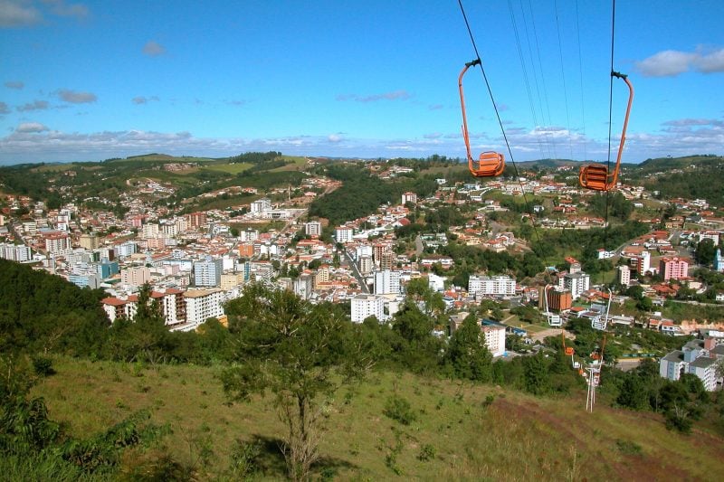 Vista do Teleferico em Serra Negra, com cabines suspensas sobre a cidade e morros verdes ao fundo sob céu azul com nuvens.