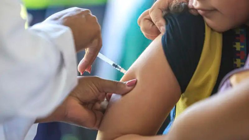 Sarampo em SP: profissional aplica vacina em uma criança, segurando a seringa no braço para imunização, com foco no momento da aplicação e alerta sobre casos no estado.