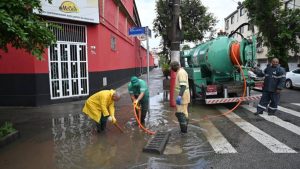 Equipe realizando drenagem e limpeza em Santos após fortes chuvas, com bombeamento e desobstrução de uma sarjeta na rua molhada em área urbana